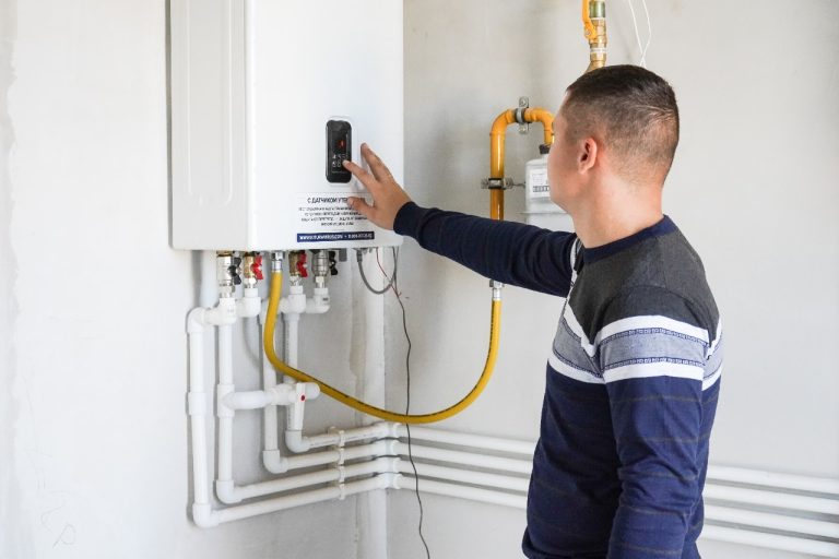 A man in a striped sweater adjusts the digital control panel of a white, wall-mounted tankless gas water heater.